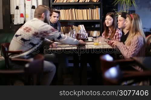 Hipster teenagers sitting at cafe table and chatting. Group of young friends relaxing after colllege in coffee shop and making pile of different mobile phones on the table. Classmates communicating after lessons in cafeteria.