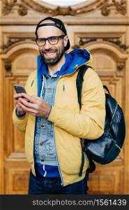 Hipster tourist in glasses, cap and yellow anorak holding backpack and smartphone having excursion in art gallery making photos being happy and delightful. People, tourism, technology concept