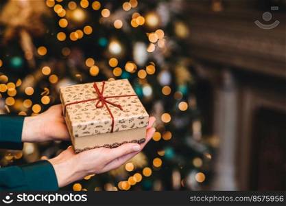 Hirizontal shot of beautiful wrapped present box in woman s hands against decorated Christmas or New Year tree. New Year background. Focus on gift box. Celebration concept.