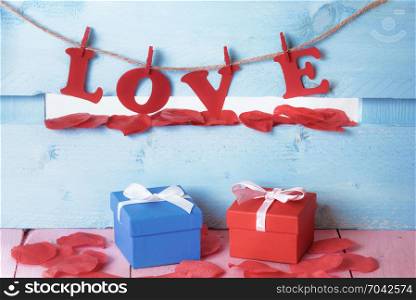 His and hers gift boxes surrounded by soap red petals and the word love spelled with red paper letters tied to a string, on a blue wooden fence.