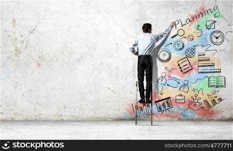His bright strategy plan . Back view of businessman standing on ladder and drawing on wall