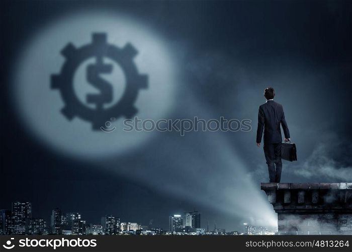 His goal is to become rich. Businessman standing with back in darkness and dollar sign in spothlight