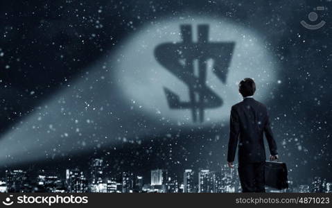 His goal is to become rich. Businessman standing with back in darkness and dollar sign in spothlight