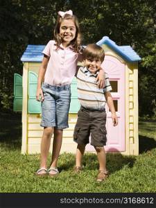 Hispanic boy and girl in front of outdoor playhouse smiling at viewer.