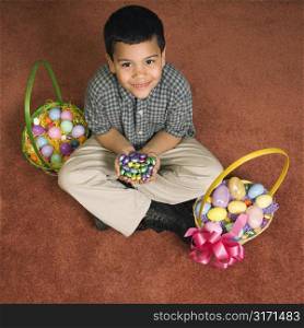 Hispanic boy sitting on floor with two Easter baskets holding chocolate candy eggs in his hands looking up at viewer smiling.