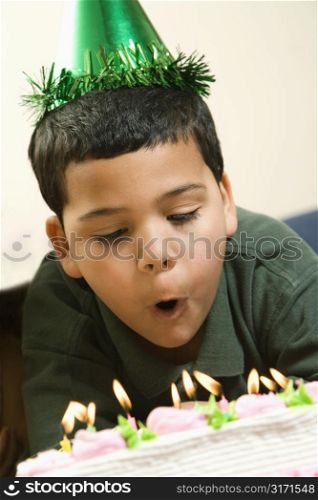 Hispanic boy wearing party hat blowing out candles on birthday cake.
