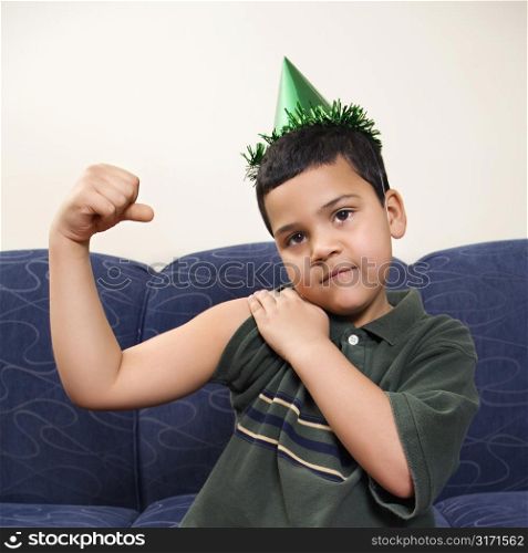 Hispanic boy wearing party hat playfully flexing arm muscle while looking at viewer.