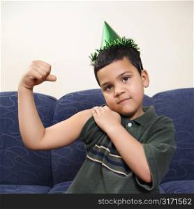 Hispanic boy wearing party hat playfully flexing arm muscle while looking at viewer.