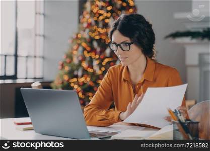 Hispanic business lady in glasses working online in office on Christmas, sitting at desk and looking on laptop screen, female employee using computer at her workplace during xmas holidays. Hispanic business lady in glasses working online in office on Christma