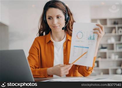Hispanic businesswoman in headphones is brainstorming online from home. Corporate internet session on laptop. Young pretty leader in orange shirt shows charts and points to the data. Remote business.. Hispanic businesswoman in headphones is brainstorming online from home. Leader shows charts.