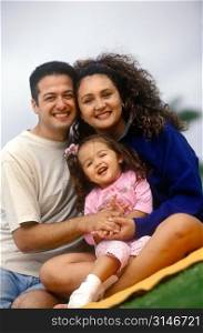 Hispanic Family Sitting On A Blanket In The Park And Smiling