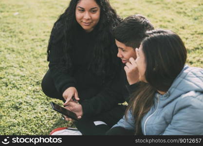 Hispanic family spending quality time together in a local park on a beautiful, sunny day. A teenage boy is sitting on the grass, holding his smartphone while looking away. His mother and sister are sitting next to him, looking at the camera and smiling. Hispanic family spending quality time together in a local park on a beautiful, sunny day. A teenage boy is sitting on the grass, holding his smartphone while looking away. His mother and sister are sitting next to him, looking at the camera and smiling.