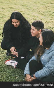 Hispanic family spending quality time together in a local park on a beautiful, sunny day. A teenage boy is sitting on the grass, holding his smartphone while looking away. His mother and sister are sitting next to him, looking at the camera and smiling.. Hispanic male teenager holding smartphone looking away while sitting on grass with Hispanic mother and sister in park on sunny day