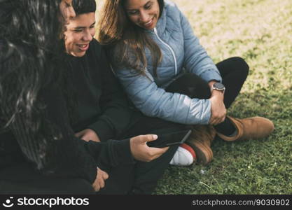Hispanic family spending quality time together in a local park on a beautiful, sunny day. A teenage boy is sitting on the grass, holding his smartphone while looking away. His mother and sister are sitting next to him, looking at the camera and smiling.. Hispanic male teenager holding smartphone looking away while sitting on grass with Hispanic mother and sister in park on sunny day