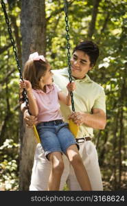 Hispanic father pushing daughter on swing and making eye contact.