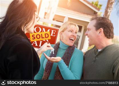Hispanic Female Real Estate Agent Handing Over New House Keys to Happy Couple In Front of Sold For Sale Real Estate Sign.