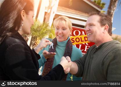 Hispanic Female Real Estate Agent Handing Over New House Keys to Happy Couple In Front of Sold For Sale Real Estate Sign.