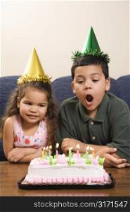 Hispanic girl and boy wearing party hats preparing to blow candles out on birthday cake.
