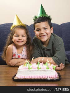 Hispanic girl and boy wearing party hats sitting in front of birthday cake smiling and looking at viewer.