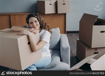 Hispanic girl relocates to new home. Lady is sitting in armchair among packed cardboard boxes. Woman is dreaming leaned to box. Happy young woman moves alone. Independence and success concept.. Hispanic girl relocates to new home. Lady is sitting in armchair among boxes and dreaming.