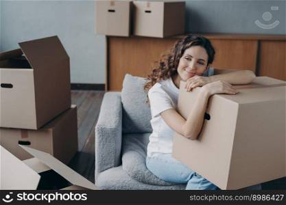 Hispanic girl relocates to new home. Lady is sitting in armchair among packed cardboard boxes. Woman is dreaming leaned to box. Happy young woman moves alone. Independence and success concept.. Hispanic girl relocates to new home. Lady is sitting in armchair among boxes and dreaming.