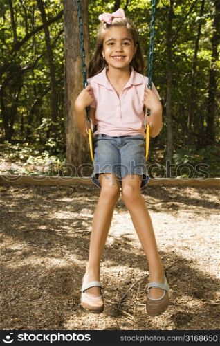 Hispanic girl sitting on playground swing smiling at viewer.