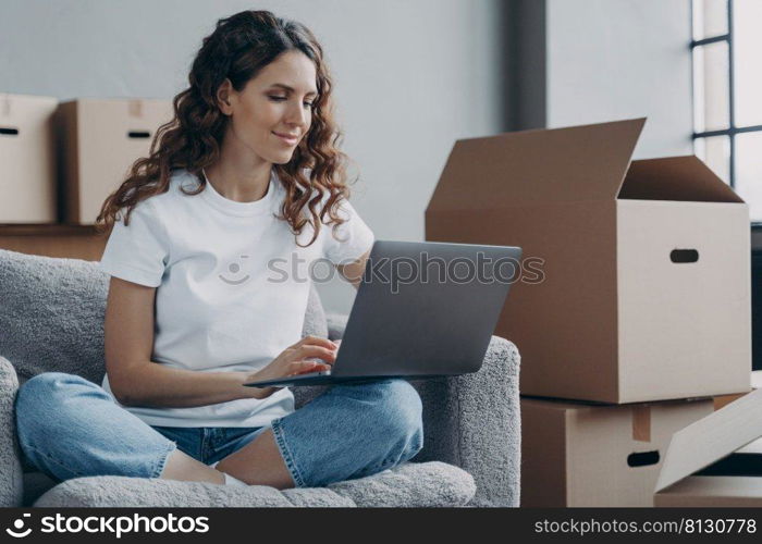 Hispanic girl sitting with laptop among cardboard boxes and smiling. Happy young woman using internet in new home. Lady moves to modern apartment. Relocation and online work concept.. Hispanic girl sitting with laptop among cardboard boxes and smiling. Relocation and online work.