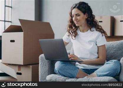 Hispanic girl sitting with laptop among cardboard boxes and smiling. Happy young woman using internet in new home. Lady moves to modern apartment. Relocation and online work concept.. Hispanic girl sitting with laptop among cardboard boxes and smiling. Relocation and online work.