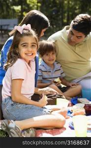 Hispanic girl smiling at viewer with family picnicking in the park.