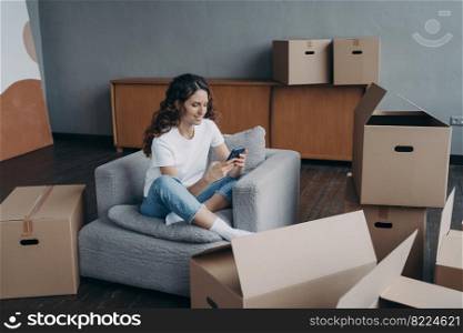 Hispanic girl uses phone app, searching moving company, shopping online for new home, sitting on armchair with carton boxes during packing things. Relocation, apartment renovation concept.. Girl uses phone app, orders moving company, shopping online for new home, sitting with carton boxes