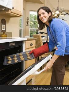 Hispanic mid adult woman putting cookies into oven and smiling at viewer.