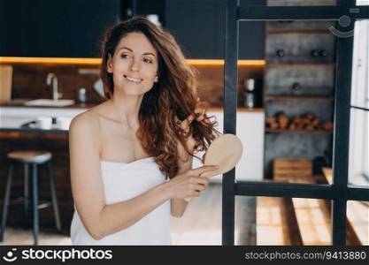 Hispanic woman brushes long curly hair. Relaxed girl in towel. Modern apartment. Home p&ering.