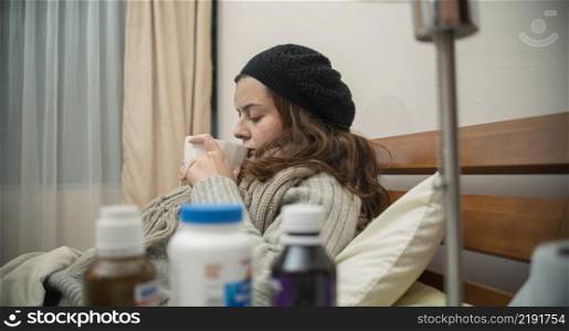 Hispanic woman lying alone in her well-dressed bed next to a table full of medicine, sick with the flu, drinking a cup of tea. Hispanic woman lying alone in her bed well wrapped up, sick with the flu, drinking a cup of tea