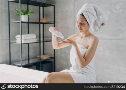 Hispanic woman using natural skincare oil in bathroom. Female wrapped in towel holding mockup bottle with moisturizing lotion sitting on bathtub. Body skin care treatment, organic cosmetics ad.. Woman holding mockup bottle with natural skincare cosmetics in bathroom. Body care, skincare, spa