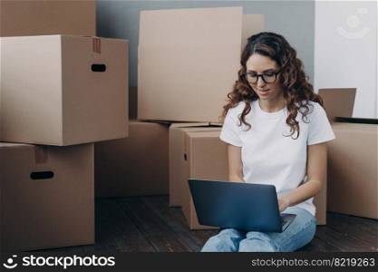 Hispanic woman with laptop searching for mover for relocation online sitting on floor with cardboard boxes. Focused female in glasses reviewing arrangements with moving company on computer.. Female review moving company document on laptop online sitting on floor with boxes for relocation