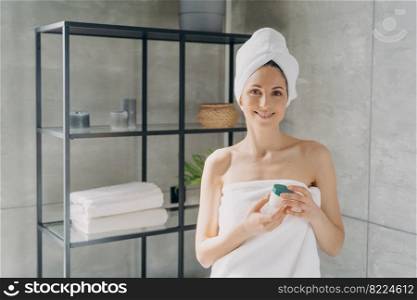 Hispanic woman wrapped in towels holding face cream jar, looking at camera in bathroom. Smiling female using anti-wrinkle treatment serum after shower. Skincare daily routine, self-care.. Hispanic woman holding face cream jar after shower in bathroom. Skincare daily routine, self-care