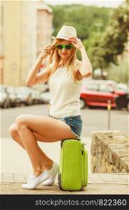 Hitch hiking and travelling. Lovely smiling cute girl sits on green suitcase luggage baggage waiting for car. Female hitch-hiker in hat travelling all around the world.. Woman traveler sits on suitcase waiting for car.