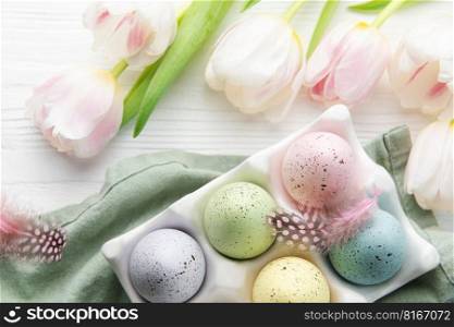 Holder  with Easter eggs painted pastel colors and pink tulips on a white wooden background. Top view. Space for text.