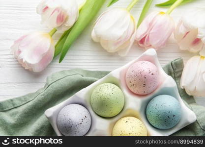 Holder  with Easter eggs painted pastel colors and pink tulips on a white wooden background. Top view. Space for text.