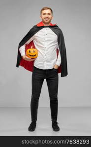 holiday and people concept - happy smiling man in halloween costume of vampire and dracula cape holding jack-o-lantern pumpkin over grey background. man in halloween costume of vampire with pumpkin