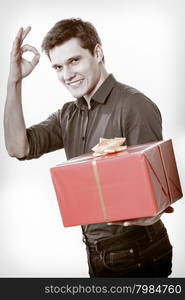 Holiday and special occasion. Young handsome man giving red present gift box with golden ribbon and showing ok okay success hand sign isolated on white. Award surprise. Studio shot.