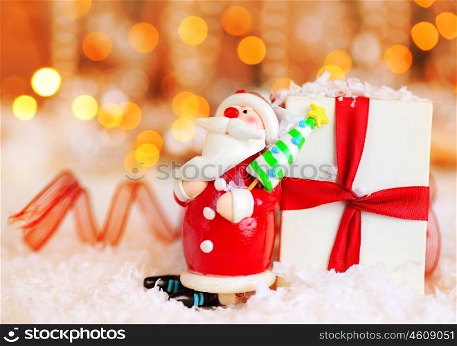 Holiday background with cute Santa Claus Christmas tree decorative ornament &amp; gift box in snow over abstract defocus lights