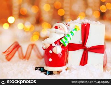 Holiday background with cute Santa Claus Christmas tree decorative ornament &amp; gift box in snow over abstract defocus lights