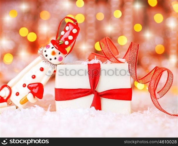 Holiday background with cute snowman Christmas tree decorative ornament &amp; gift box in snow over abstract defocus lights