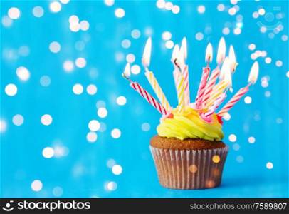 holiday, celebration, greeting and party concept - birthday cupcake with buttercream frosting and many burning candles over festive lights on blue background. birthday cupcake with many burning candles
