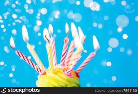 holiday, celebration, greeting and party concept - birthday cupcake with buttercream frosting and many burning candles over festive lights on blue background. birthday cupcake with many burning candles