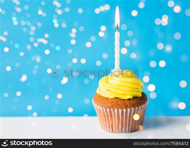 holiday, celebration, greeting and party concept - birthday cupcake with yellow buttercream frosting and one burning candle over festive lights on blue background. birthday cupcake with one burning candle