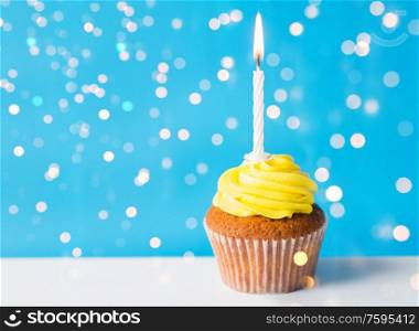 holiday, celebration, greeting and party concept - birthday cupcake with yellow buttercream frosting and one burning candle over festive lights on blue background. birthday cupcake with one burning candle