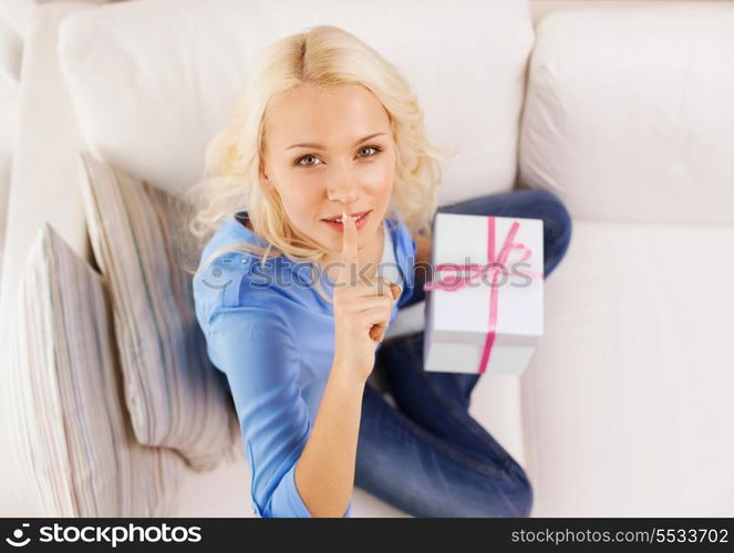 holiday, celebration, home and birthday concept - smiling young woman with gift box and making shh gesture at home