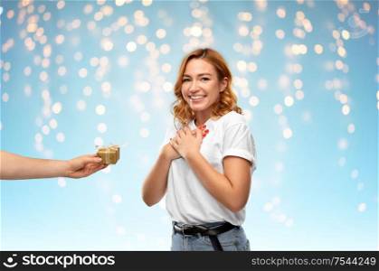 holiday, christmas and valentine&rsquo;s day concept - happy woman in white t-shirt receiving little gift box from man over holidays lights on blue background. happy couple in white t-shirts with christmas gift